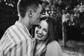 At Lac Vert de Canéjan in Gironde, France, the fiancé affectionately kisses his future wife on the temple, making her laugh during their serene and natural outdoor engagement portrait session.