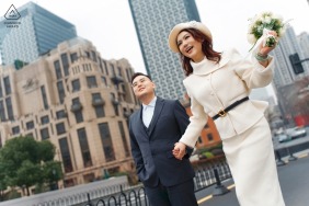 She beams with happiness and lifts her bouquet high as the couple strolls hand in hand through the vibrant city streets along the Bund in Shanghai, China, for their engagement.
