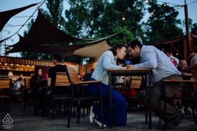 The couple sits closely at an outdoor bar in Budapest, Hungary, leaning in to share a drink under glowing string lights, surrounded by the energy of an inviting evening scene.