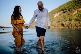 As golden sunset light bathes the shallow river in Szigetcsúcs, Hungary, a couple walks hand in hand, surrounded by lush green hills that add a poetic touch to their riverside stroll.