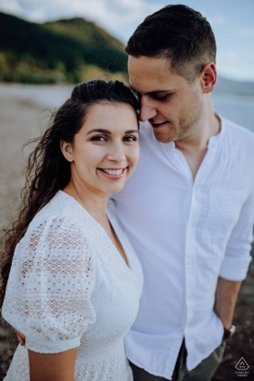 Next to the peaceful lakeshore in Szigetcsúcs, Hungary, the woman smiles warmly at the camera while her partner gently leans in, their closeness mirrored in the calm water beside them.