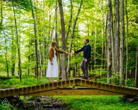 A couple enjoys the tranquility of a bridge in the woods in the Catskills, New York, their engagement portrait set amidst the lush greenery and natural beauty of the area.