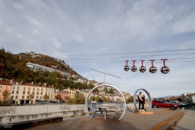 The couple stands together near the iconic gondola bubbles on wires in Grenoble, France, their connection highlighted against the playful, urban setting of this city venue known for its unique cable cars.