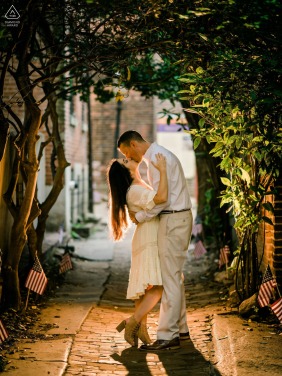 A couple shares a tender kiss in a quiet, tree-lined alley in Philadelphia, warm golden hour light filtering through the leaves and casting a soft glow over the romantic engagement portrait.