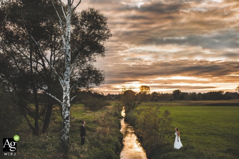 Grotniki, Lodzkie, Poland couple posing for a wedding picture over a small river with big trees and clouds