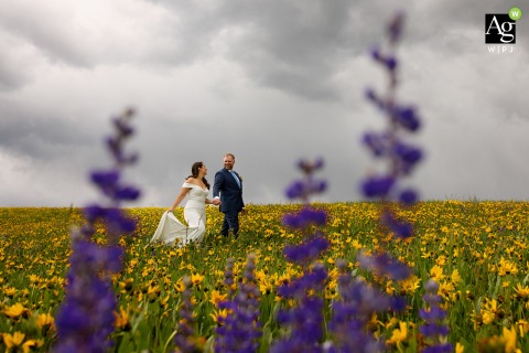 At a private residence in Steamboat Springs, Colorado, a bride and groom pose for a wedding day portrait, walking hand in hand through a picturesque field adorned with daisies and lupine wildflowers.