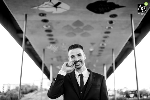Beneath a bridge in the Edmonton river valley, the groom pauses for a portrait, thoughtfully twirling his moustache, with the urban landscape framing his unique style.