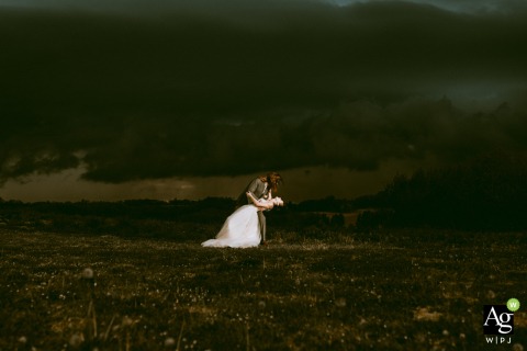 Storm clouds gather above Domaine des Gaillardoux in Lot, France, as the bride and groom stand close and dip during their photography session, their attire in striking contrast to the dramatic sky.