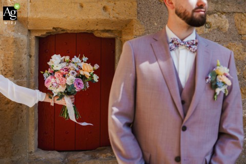 Against a rustic stone backdrop at Porte des Allemands in Metz, a hand offers a colorful bouquet to the groom in his soft pink suit, with an old red door adding warmth and charm.