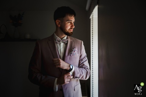 During preparations in Moselle, an elegantly dressed man in a pale pink suit and floral bowtie adjusts his watch by a softly lit window, his thoughtful expression enhanced by intimate, gentle light.