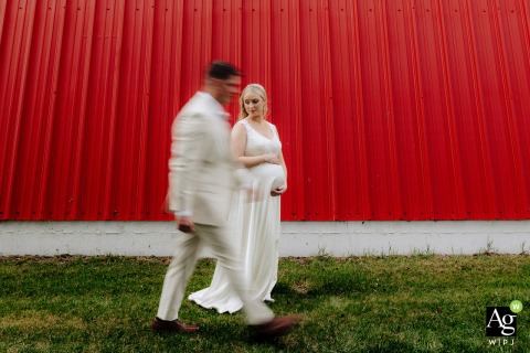 The groom walks past his pregnant bride against the striking backdrop of a red barn wall in Edmonton, Alberta, creating a unique and meaningful rural wedding day portrait.