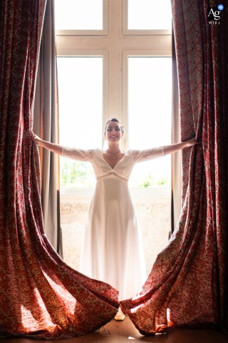Symmetrical vertical shot of the bride holding drapes open at a window at Domaine de la Dame Blanche, Gironde, France.