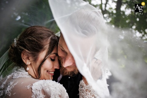 A couple in a close embrace, smiling under the bride's veil amidst trees, during their wedding portrait session at Le Grand Hotel, Le Pont, Switzerland.