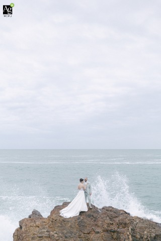 Perched on jagged rocks overlooking the ocean in Vendée, France, the bride and groom pose for a dramatic portrait as a large wave crashes behind them following their ceremony.