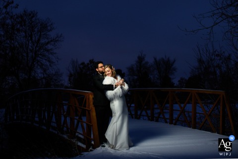 Bride and groom embrace lovingly on bridge at Wellshire Event Center during enchanting twilight wedding.