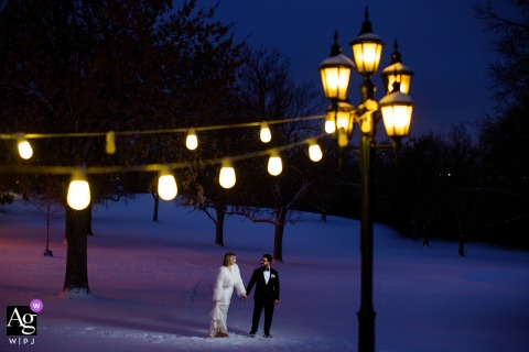 Bride and groom walk hand-in-hand through snowy landscape at Wellshire Event Center during twilight.