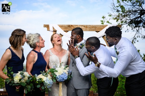 The wedding party gathers around the groom at Christies of Genesee in Colorado, leaning in to admire his new wedding ring during a lighthearted group portrait session.