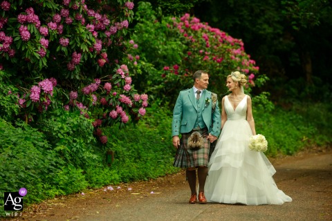 Bride and groom walk by colorful flowers at Balbirnie House, Glenrothes, Scotland, smiling together.