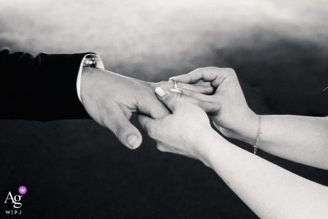 A black and white detail shot capturing the emotional scene of the ring exchange as the bride slides the ring onto the groom's finger during the ceremony at Ranch des Rochettes, Cezeriat, Ain, France.