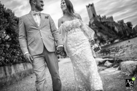 A timeless black and white portrait features the newlyweds walking along the lakeside promenade in Malcesine, Italy, with the iconic silhouette of the castle rising behind them.