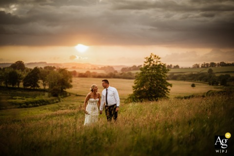 An artistic wedding portrait from Dorset, England, featuring the couple holding hands in a sun-drenched, dry grass field in Chedington, with the low sun behind them.