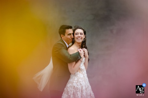 Soft, pastel-toned wedding portrait of the bride and groom at Denver Art Museum, Denver, CO, viewed through florals as he holds her from behind.