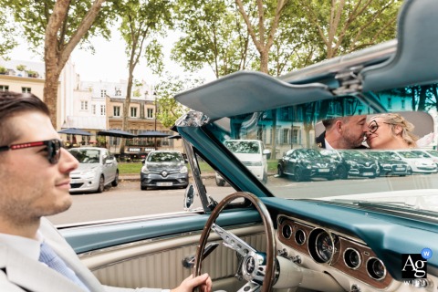 Versailles Kiss: Bride and Groom Reflected in Convertible Mirror as Driver Focuses Bride and groom kissing in the reflection of a convertible car's mirror in Versailles, Yvelines, as the driver focuses on the city roadway.