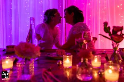 La Bullerie winery provides the backdrop as the newlyweds lean intimately toward each other at a dinner table, with a colorful, artistic glow of lights creating a vibrant portrait behind them on their wedding day.
