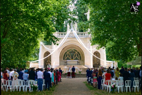 An artistic scene setter of the church, captured before the outdoor ceremony, features rows of empty wooden chairs facing a simple altar beneath a large, spreading tree on the sun-dappled grounds.