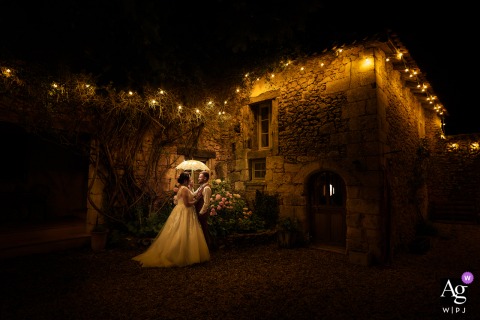Dordogne, France, bride and groom stand bathed in the warm, artistic glow of the lights illuminating their magnificent wedding estate on their special day.