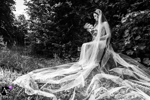 In a garden setting, the bride is seated on a bench with her veil draped elegantly around her as she holds her floral bouquet, creating an artistic portrait on her wedding day.