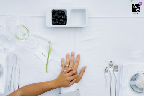 A couple's hands rest gently on a white-linened table in Amares, the bride's atop the groom's, surrounded by the subtle geometry of fine flatware.