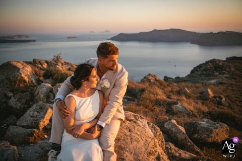 Romantic Sunset Portrait Of Couple On Rocks In Santorini Greece With Dramatic Sea Backdrop In Santorini, Greece, a couple poses on the rocks at sunset, where a warm glow illuminates their faces against the dramatic backdrop of the sea.
