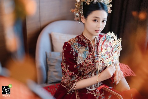 Adorned in traditional Chinese attire and an elaborate headdress, the bride sits gracefully with an ornate fan at her home in Shanghai, embodying a classic cultural aesthetic.