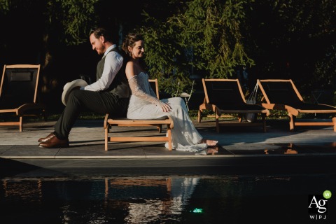 The Couple Sits Quietly Back To Back At The Edge Of A Swimming Pool In Meuse Seated back-to-back at the edge of a swimming pool, the couple sits quietly at the bride's family home in Meuse as the cocktail hour concludes.