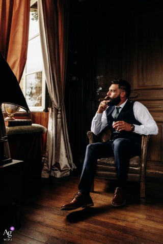 Château de la Croix — The groom sits alone by a window indoors, dressed and ready for the day, pausing reflectively at the end of getting ready in a wedding portrait.