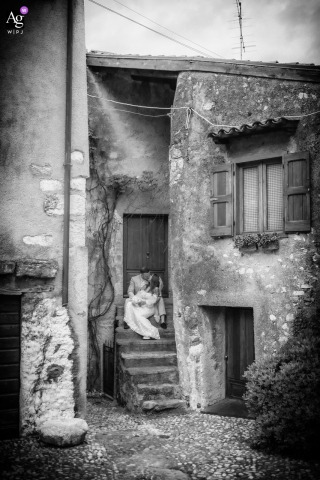Malcesine, Lake Garda, Italy — The bride and groom sit closely together on the steps of a picturesque, quintessential Malcesine street, sharing a tender and intimate session in this romantic wedding portrait.