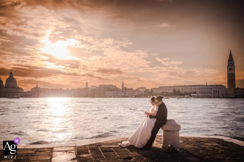 Venice, Italy — The bride and groom tenderly admire their wedding rings as the Venice skyline glows at sunset in the background, creating a romantic portrait that evokes the look of a vintage painting.