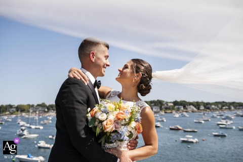 Crocker Park, Marblehead, Massachusetts — The couple smiles and embraces in front of the harbor, the bride’s veil blowing in the wind with a sea of moored boats creating a picturesque wedding portrait backdrop.