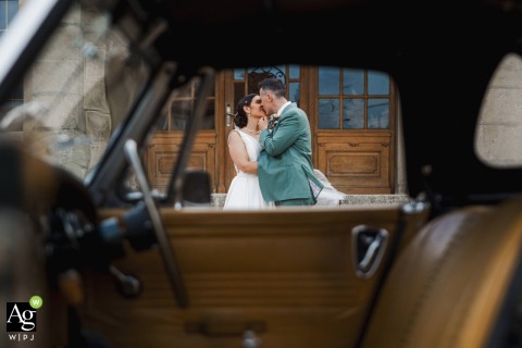 Framed through the open window of a vintage car, the newlyweds share an intimate kiss in front of a stone building at the Petit Château de Seicheprey.