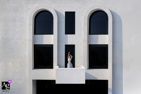 Fujian, China, hotel — The bride stands alone on the balcony, captured from afar in a symmetrically framed solo portrait that highlights the architectural building and her serene presence.