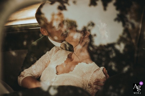 Chateau de Planchevienne — Inside a car, the couple shares a kiss that is captured through the side window glass, which reflects surrounding trees, creating a vintage-inspired and romantic wedding portrait.