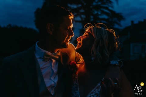 Château de Contremoret — The couple faces each other, smiling and embracing during a nighttime kiss, softly illuminated by a warm glow close to and behind them, creating a romantic and intimate wedding portrait.