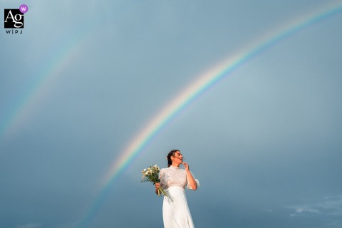 Fort la Prée, Île de Ré, France — In a minimalist wedding portrait, the bride is symmetrically framed and small at the bottom of the image, with a vast sky and double rainbow arching overhead for a striking and dreamy effect.