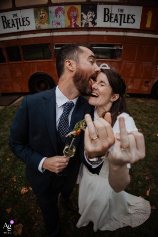 Domaine du Marchais Bouchet, France — In front of a Beatles bus, the newlyweds humorously display their ring fingers as the groom playfully licks the bride’s face, captured in a high-angle shot with a wide-angle lens for a fun wedding portrait.