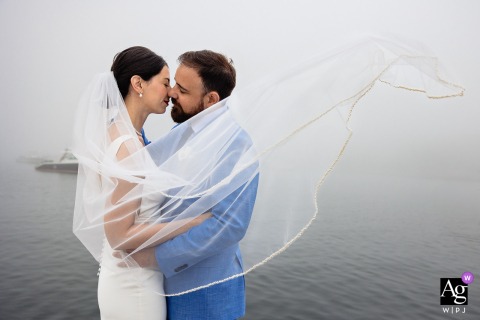 Mount Desert Island, Maine — The bride and groom kiss in the fog for a wedding portrait, her veil flowing in the wind and the sea stretching behind them, creating a dreamy and romantic coastal image.