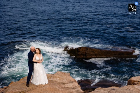 Acadia National Park, Maine — The bride and groom cuddle low in the frame on the cliffs during elopement portraits, with minimal composition and the sea stretching behind them in a high-angle shot.