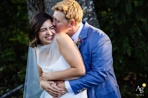 Houlton, Maine — The bride bursts with joy as she laughs during wedding portraits, her expression filling the frame while the groom stands behind her, affectionately kissing her cheek.