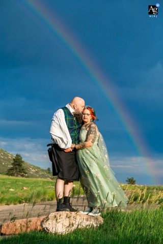 Carter Lake, Loveland, CO, USA — The couple poses perched together on a small rock amid green grass, with a vibrant rainbow arching in the background for a romantic and colorful wedding portrait.