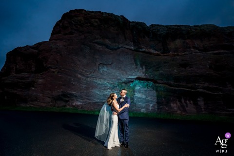 The Fort, Morrison, CO, USA — The couple poses in front of a cliff illuminated blue by the photographer at dusk, creating a dramatic and artistic wedding portrait at the historic venue.